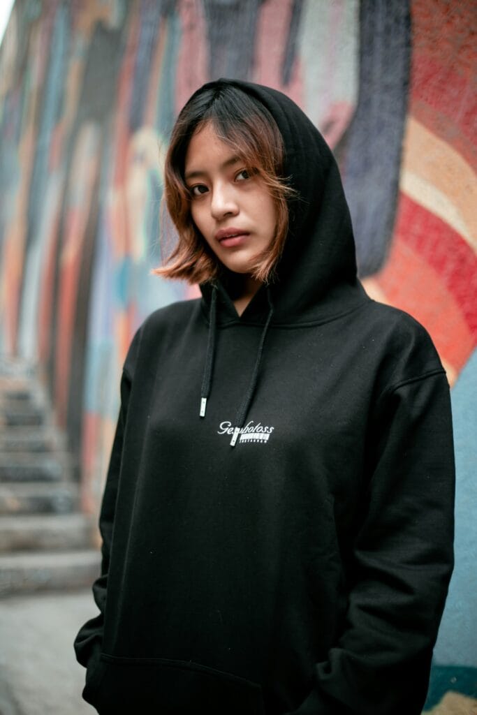 Person in a black hoodie stands in front of a colorful graffiti wall, looking at the camera.
