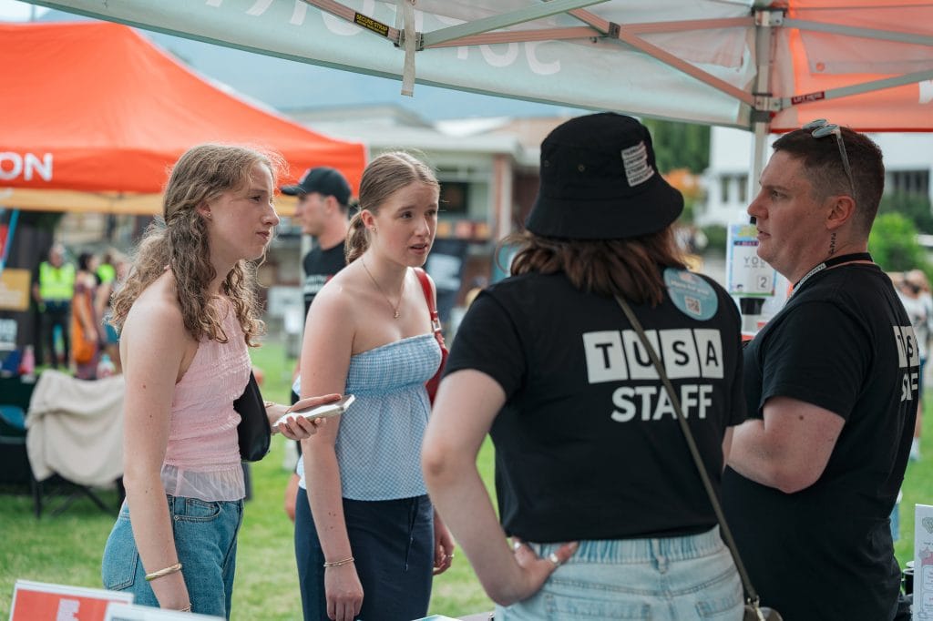 Two young women speak with two staff members at an outdoor event booth under a tent. One staff member’s shirt reads “TUSA STAFF.” Other booths and people are visible in the background.