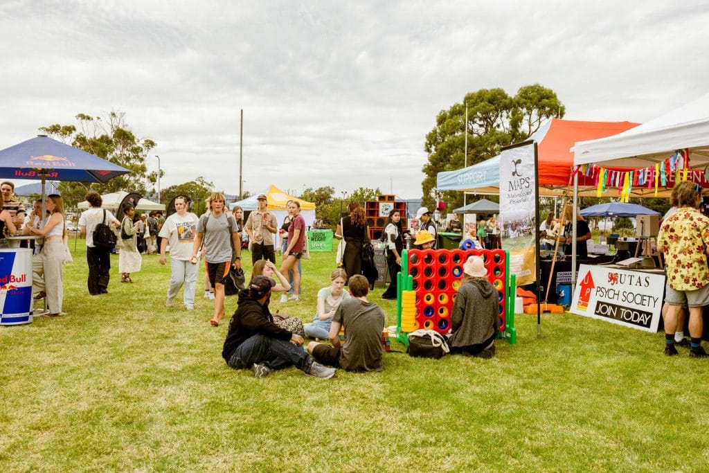 People gather on a grassy field with tents, booths, and a large Connect Four game at an outdoor event or festival.