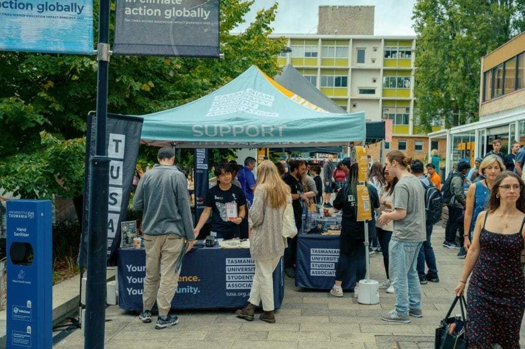 Students gather around informational booths under a tent labeled “SUPPORT” at an outdoor university event, with signs and banners visible in the background.