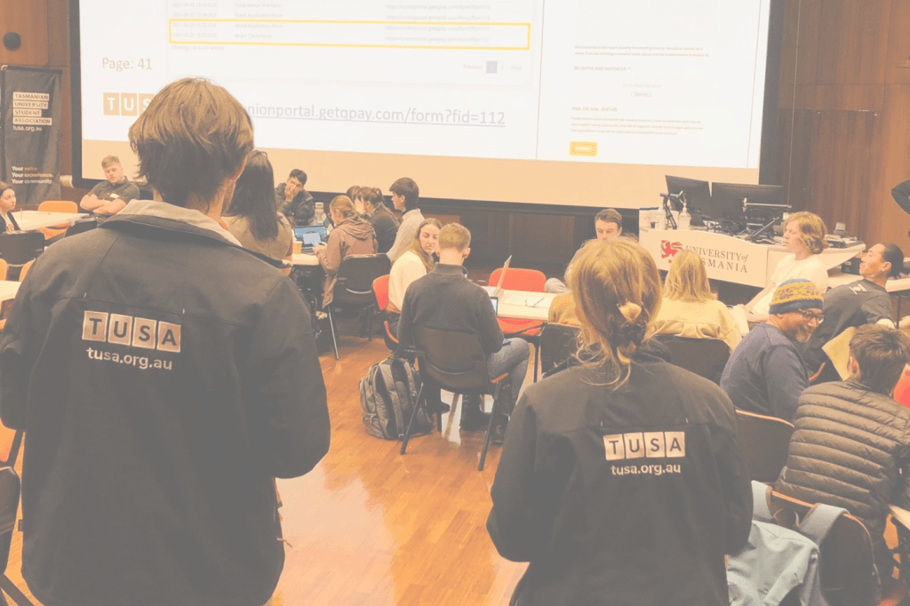 Students sit and work in groups in a university lecture hall; two people in TUSA jackets stand in the foreground, with a presentation projected on the screen.