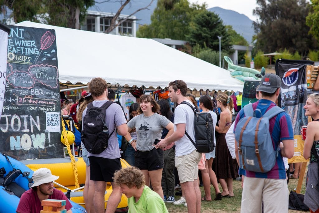 People gather outdoors at a festival or fair with booths, inflatable rafts, and informational signs under a white tent. Some are talking while others browse displays.