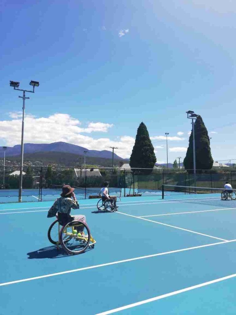 Three people in wheelchairs play tennis on a bright blue outdoor court, with mountains and trees visible in the background under a clear sky.