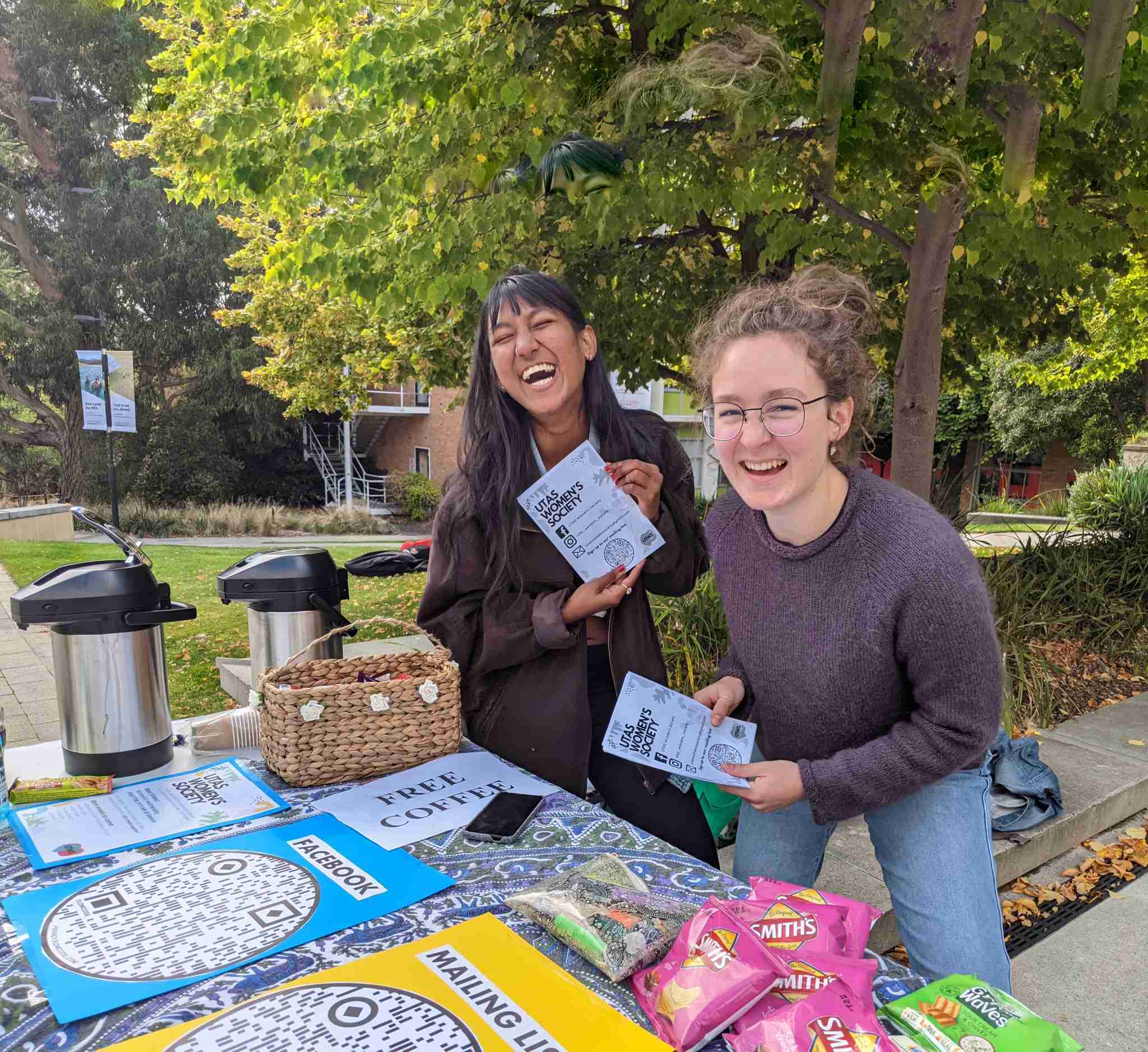 Two people stand at an outdoor table with flyers, snacks, and hot drinks, smiling at the camera under leafy trees on a campus.