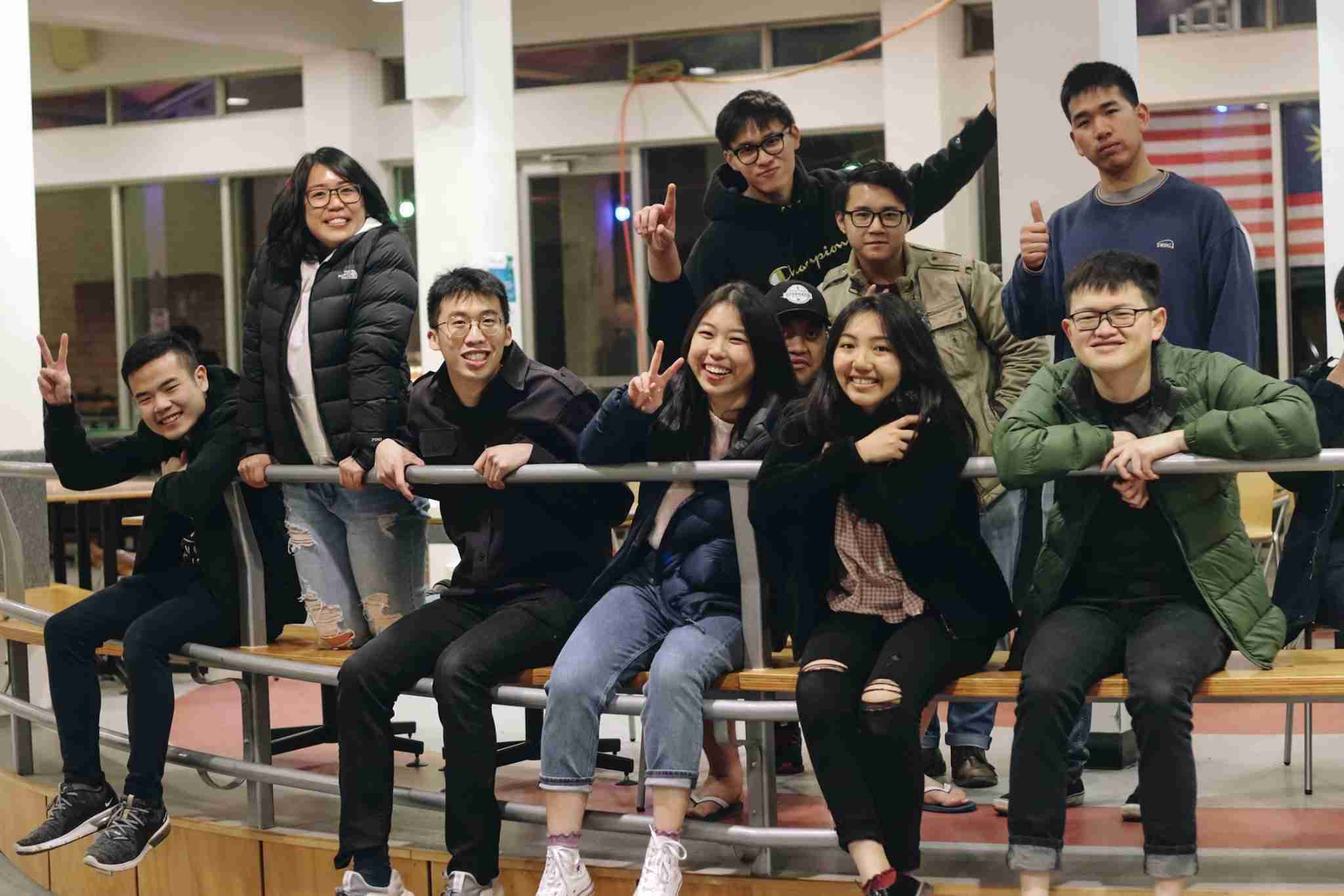 Ten young adults pose together indoors, some sitting and some standing behind a metal railing, smiling and making peace signs.