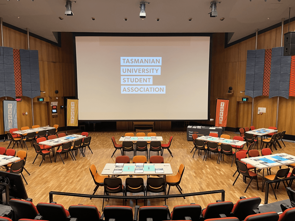 A large lecture hall set up with tables and chairs, displaying a screen that reads "Tasmanian University Student Association.