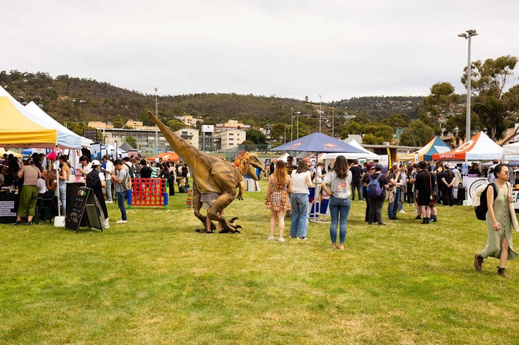 People gather at an outdoor market with colorful tents; a large dinosaur costume or model is featured in the center of the event.