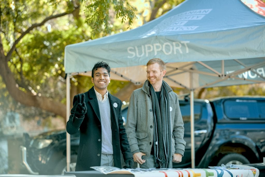 Two men in coats stand outdoors under a tent labeled "SUPPORT," smiling toward the camera, with a pickup truck and trees in the background.