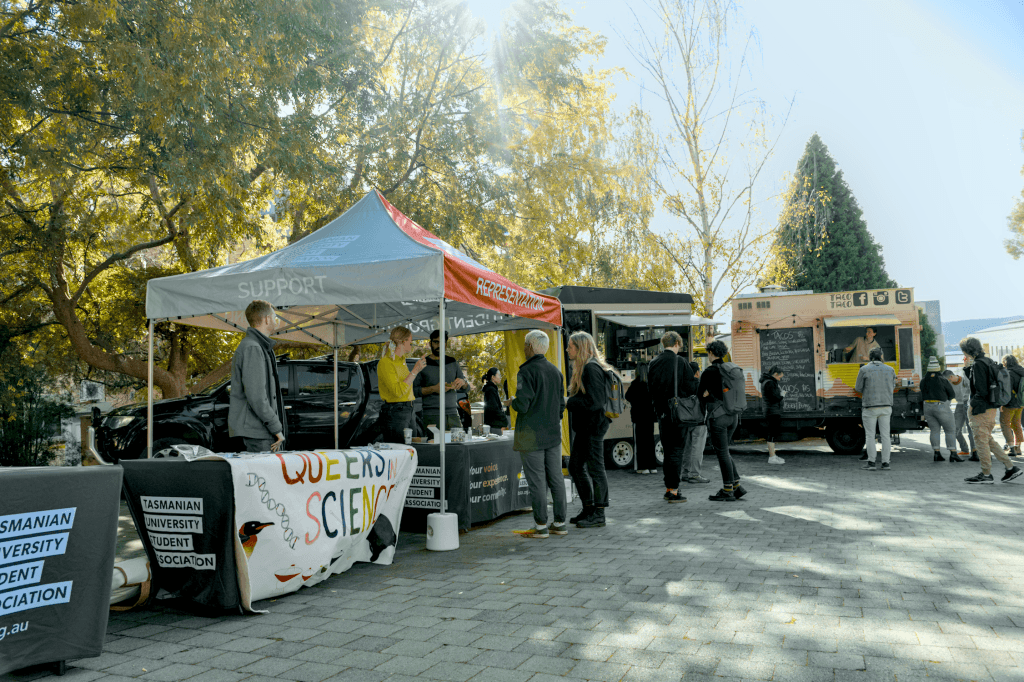 People gather at outdoor university booths and food trucks. Tables display banners, one reading "Queer Science," under trees and clear skies.