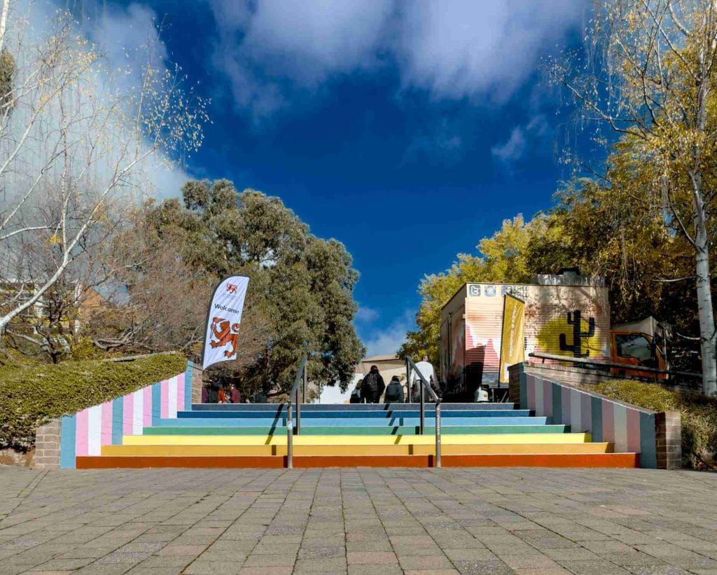 A set of colorful striped stairs leads up to a plaza with trees, a mural, and a flag on a clear day with blue sky and scattered clouds.