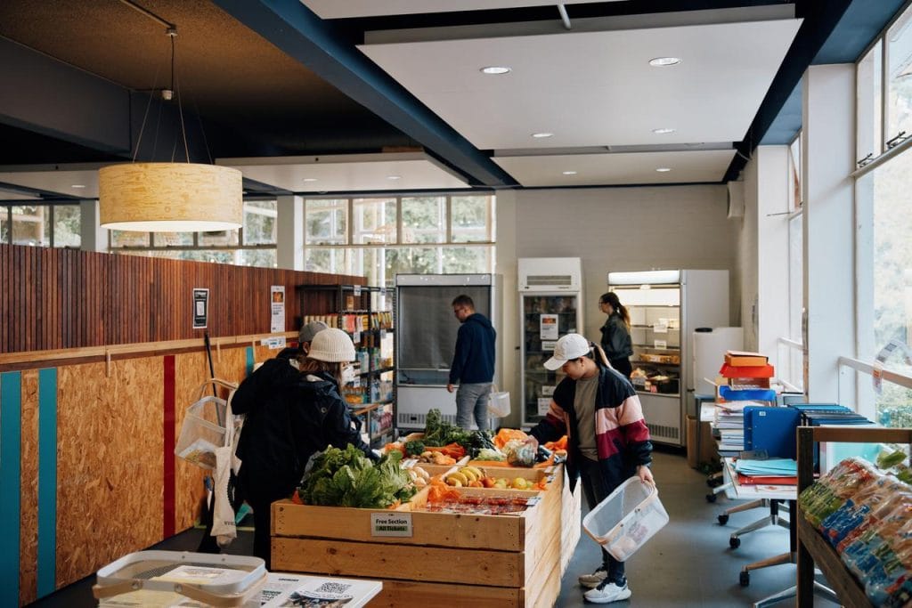 People shop for fresh produce and other groceries in a small market with wooden crates, shelves, and natural light from large windows.