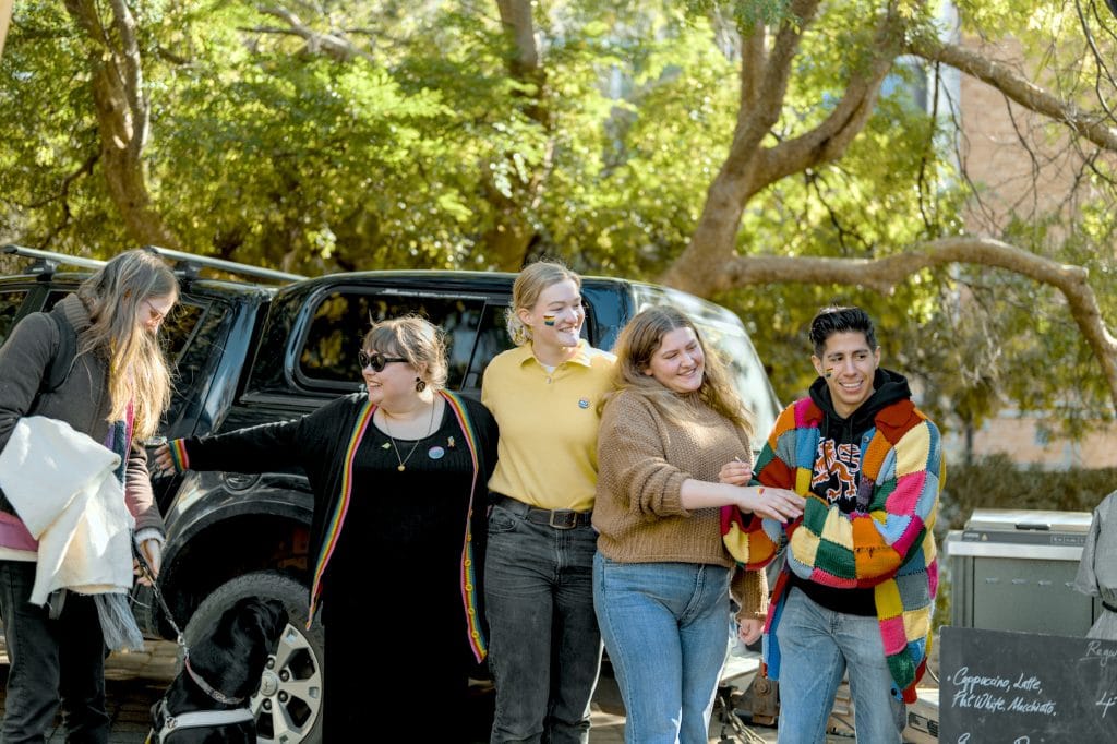 Five people stand together outdoors near a black SUV, smiling and interacting, with trees in the background and a dog partially visible on the left.