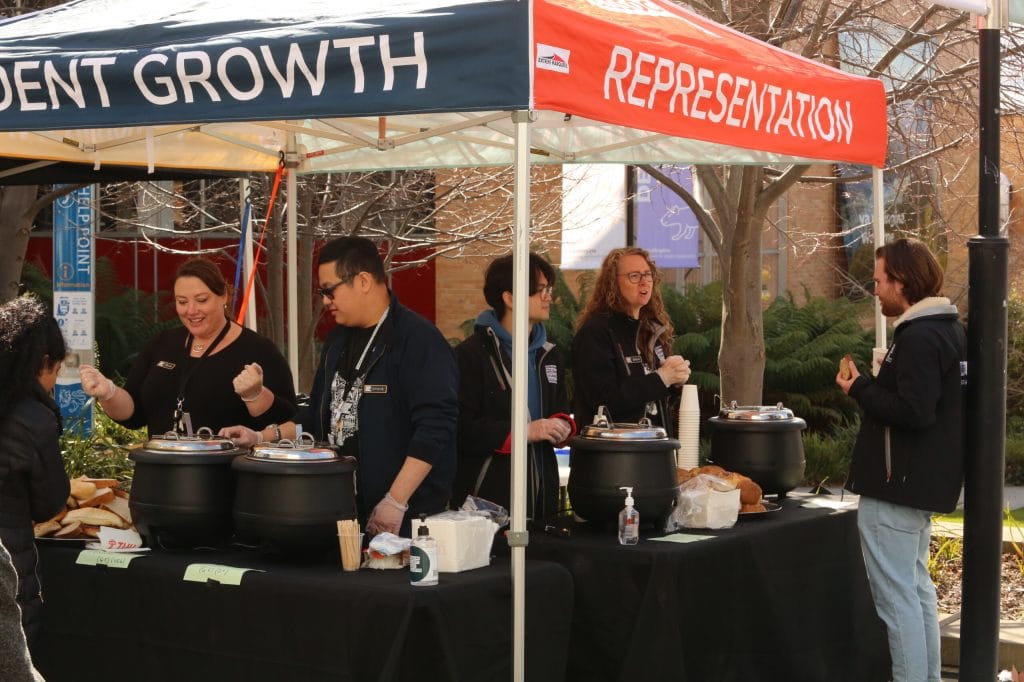 People serve food and interact under a tent labeled "STUDENT GROWTH" and "REPRESENTATION" at an outdoor event. Tables hold soup bowls, bread, and utensils.
