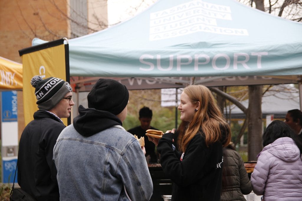 Three people stand and converse while holding food containers in front of a tent labeled "Tasmanian University Student Association SUPPORT." Other people are visible nearby.