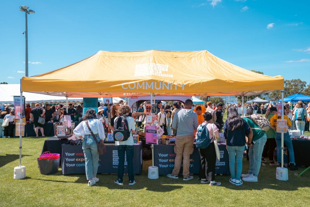 People gather around tables under a large yellow community tent at an outdoor event on a sunny day.