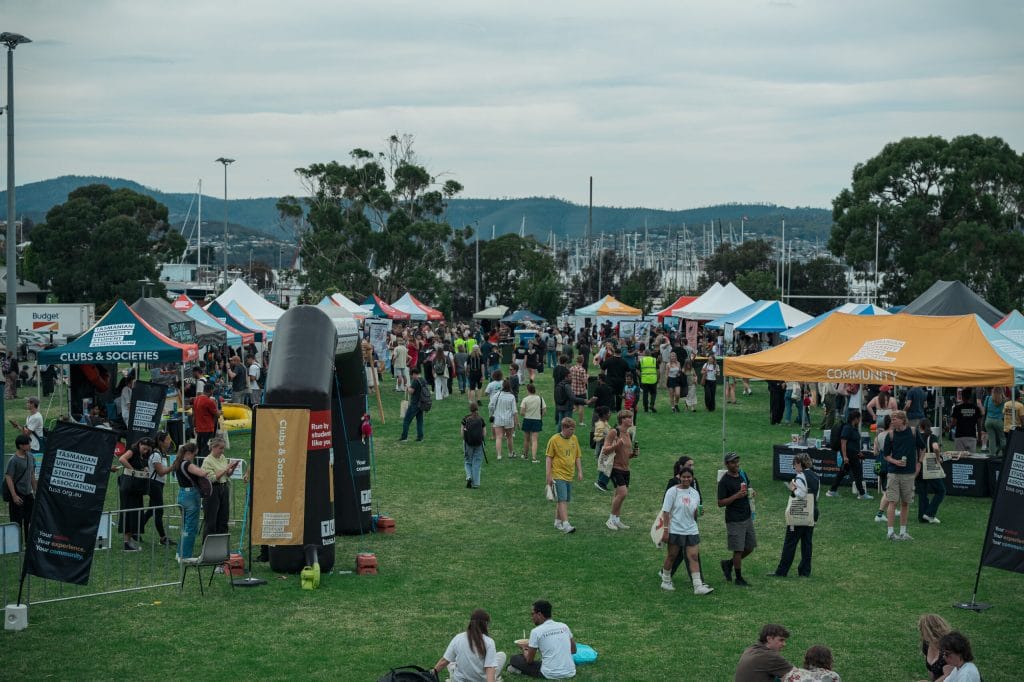A large outdoor event with people gathered among various colorful tents and information booths on a grassy field. Trees and mountains are visible in the background.