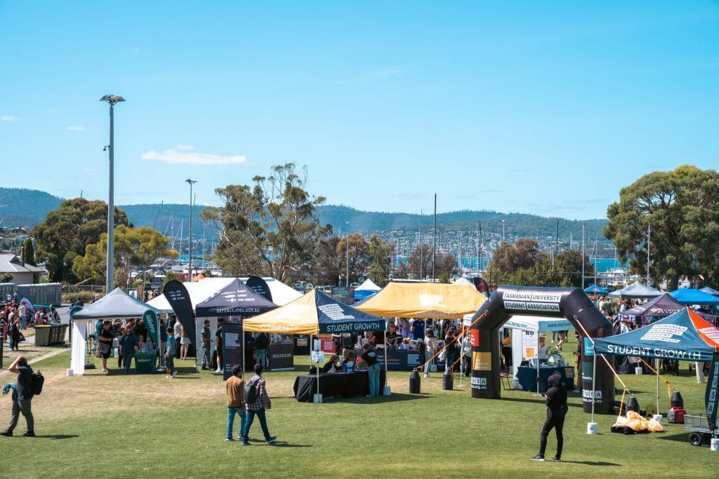 People walk among informational booths and tents set up on a grassy field during an outdoor event, with trees and boats in the background.