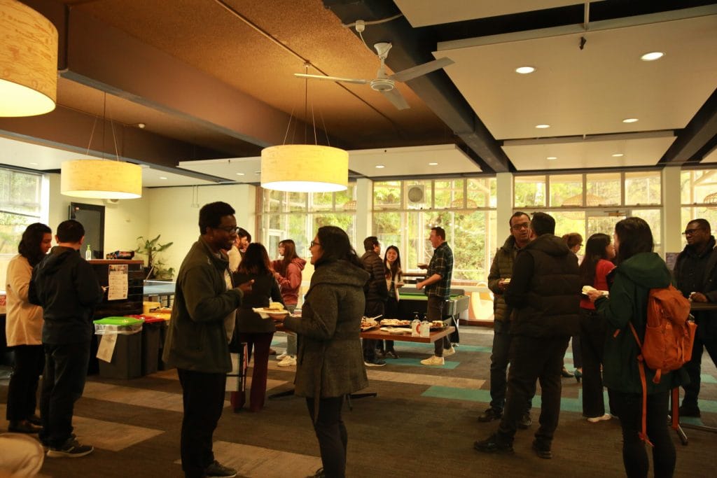 A group of people stand and converse in a well-lit room with tables of food and drinks, suggesting a social or networking event.