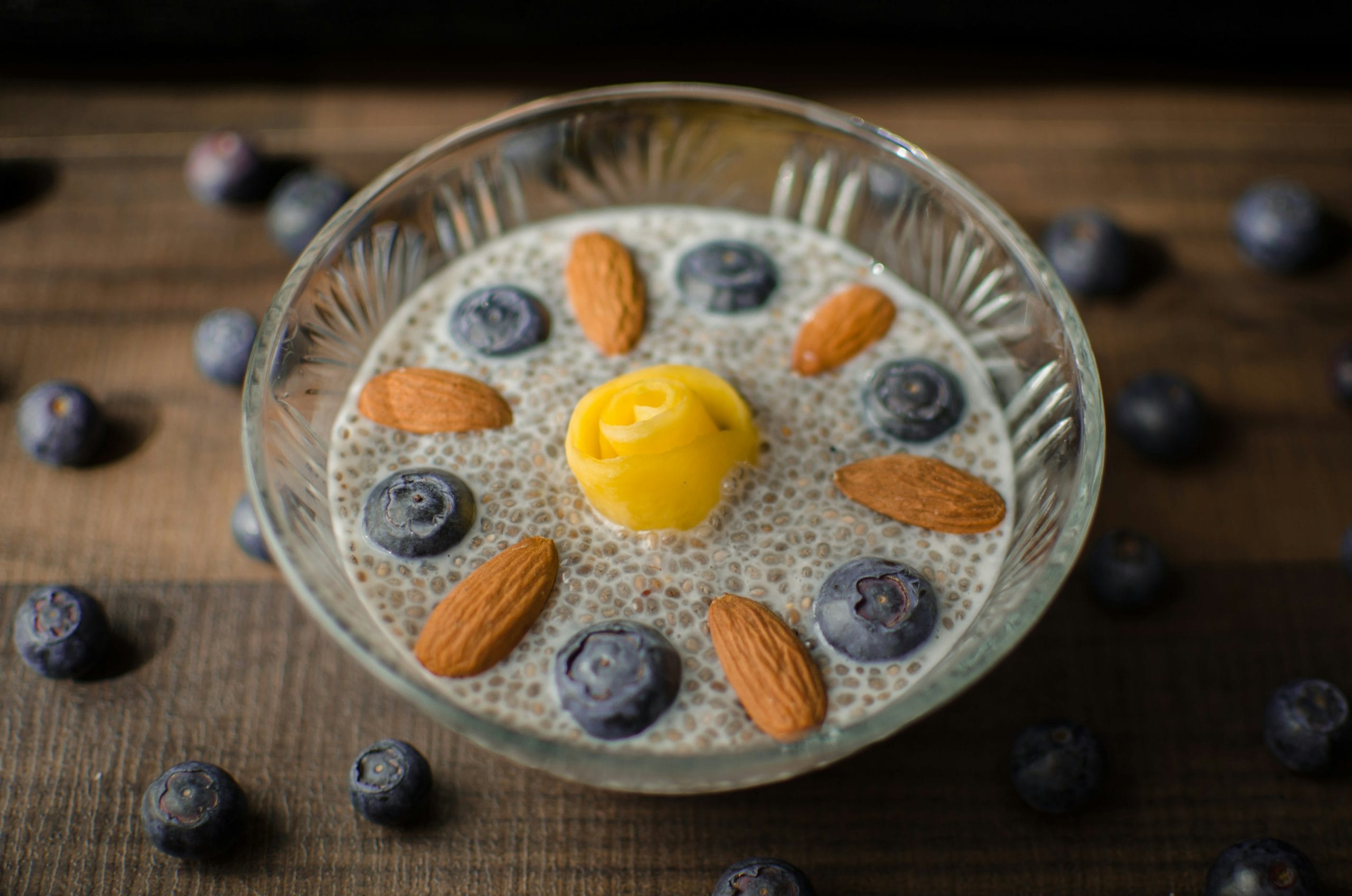 A glass bowl of chia pudding topped with blueberries, almonds, and a yellow rose-shaped fruit, surrounded by scattered blueberries on a wooden surface.