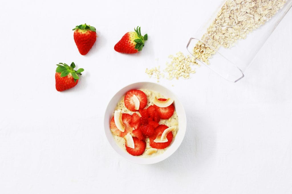 A bowl of oatmeal topped with sliced strawberries and banana, next to whole strawberries and spilled oats on a white surface.