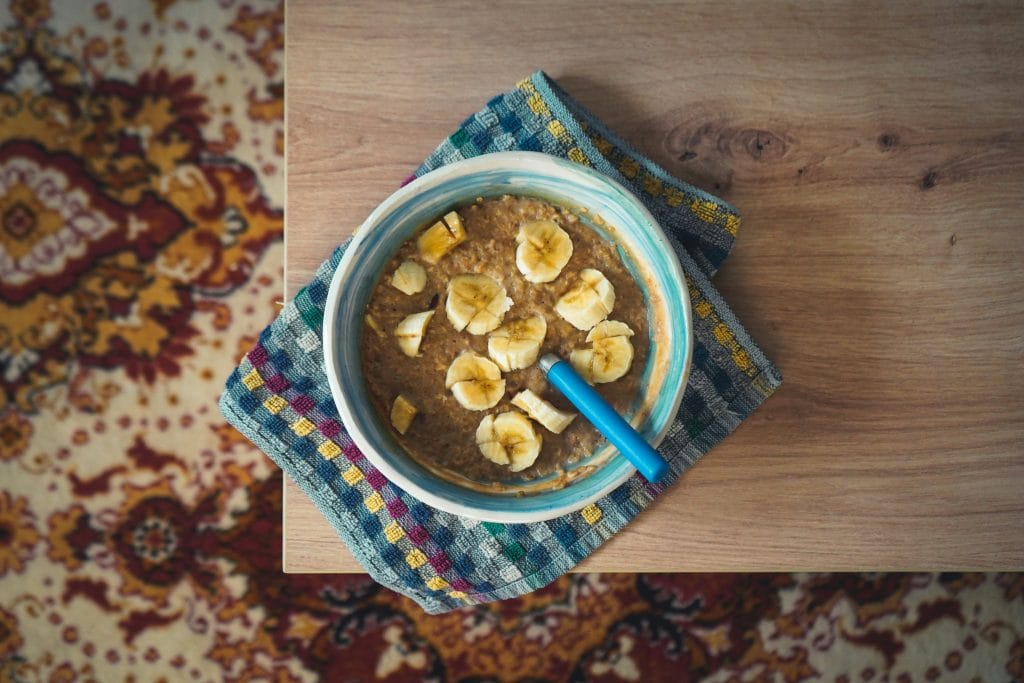A bowl of oatmeal topped with banana slices and a blue spoon sits on a checkered cloth on a wooden table next to a patterned carpet.