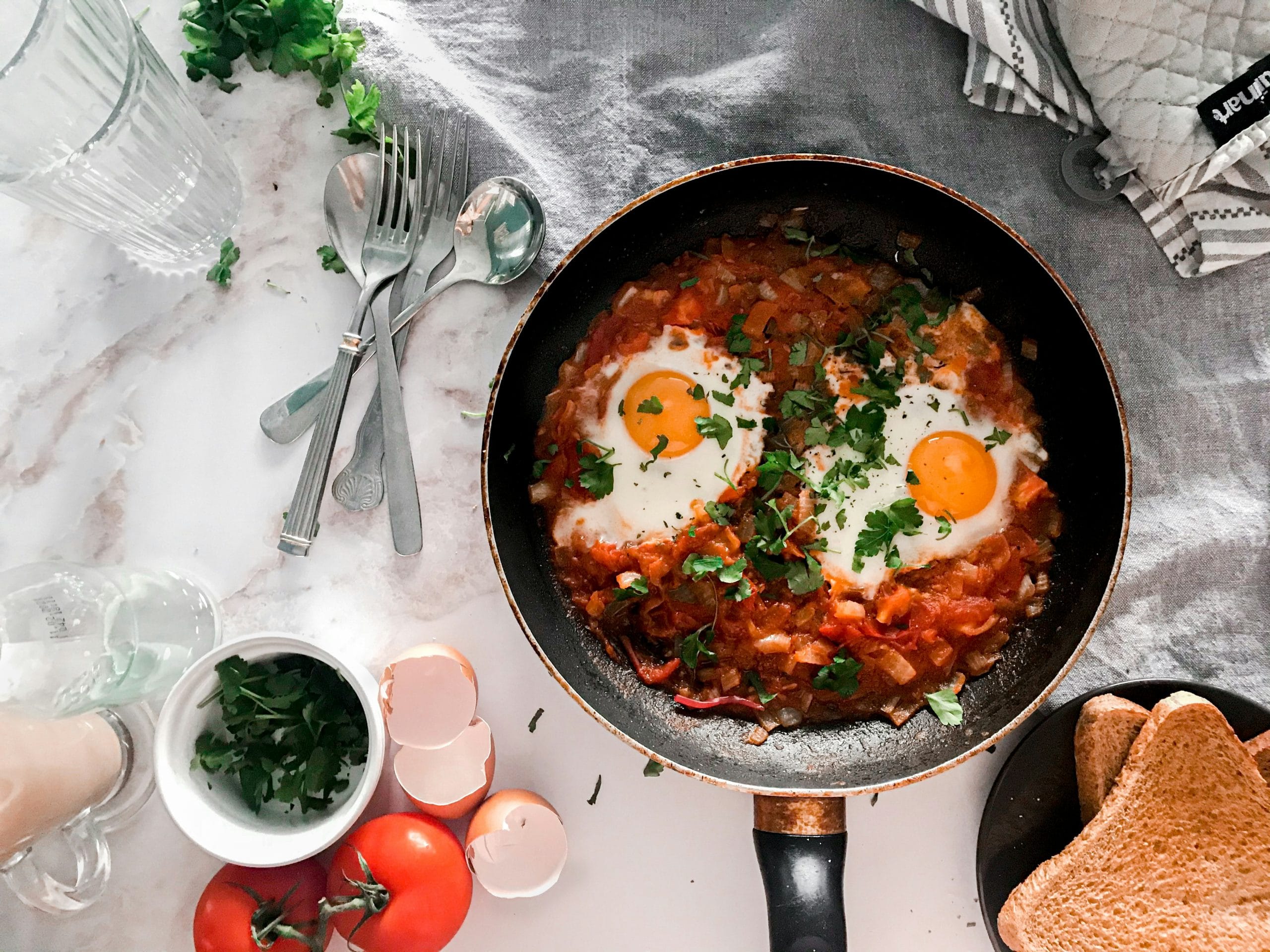 A pan with shakshuka topped with two sunny-side-up eggs and chopped herbs on a table set with toast, fresh tomatoes, utensils, a glass of water, and eggshells.
