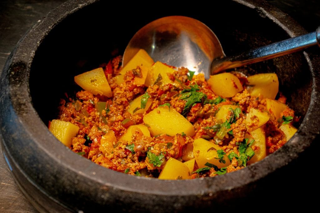 A close-up view of a black pot filled with cooked potatoes, ground meat, and herbs, with a metal serving spoon inside.