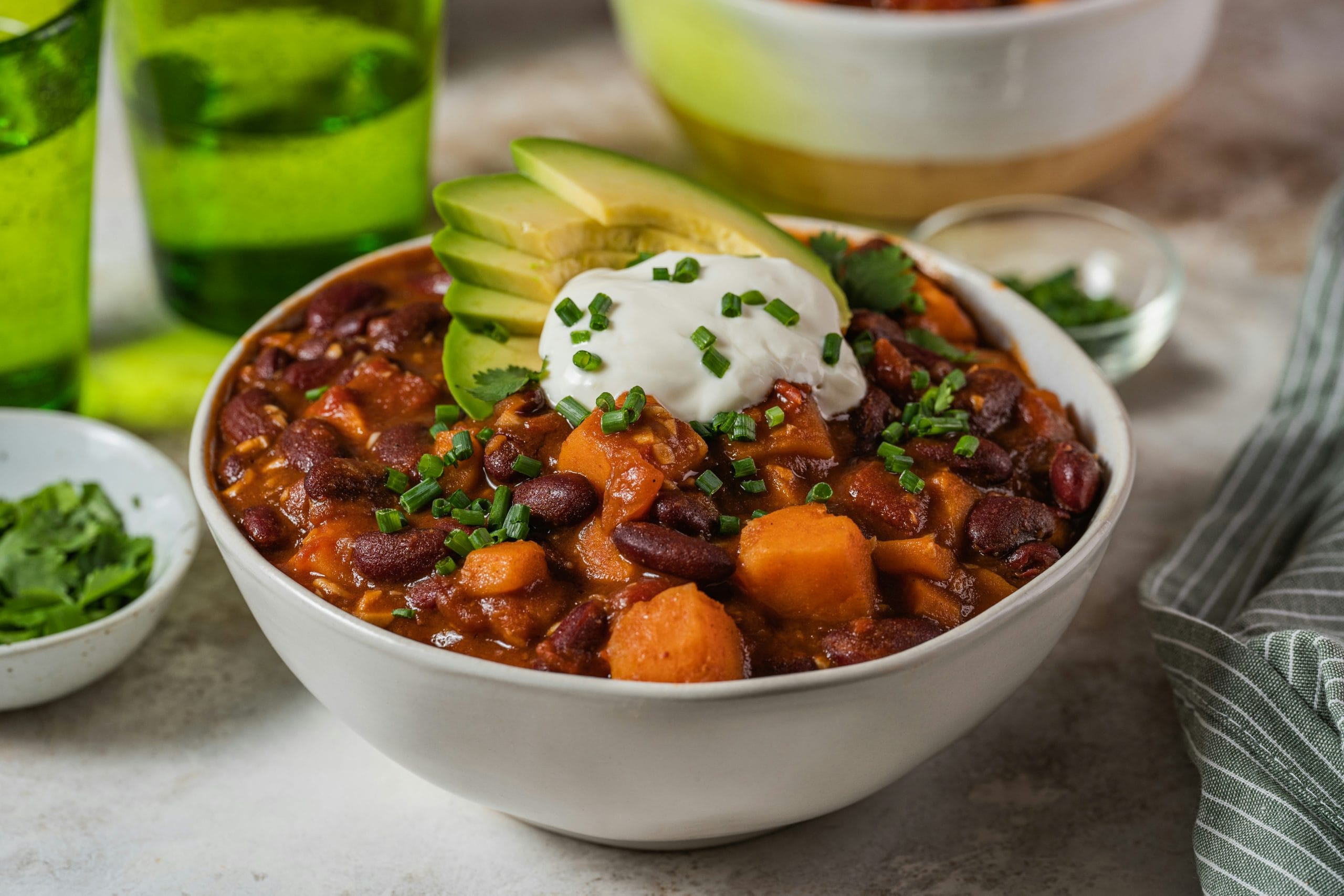 A bowl of chili with kidney beans, sweet potatoes, topped with sliced avocado, sour cream, and chopped chives, with green glasses and a striped napkin nearby.