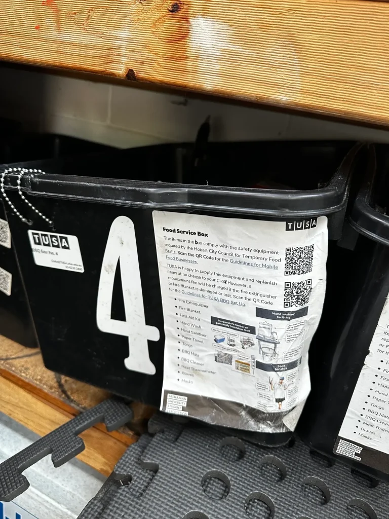 A black plastic bin labeled "4" with a TUSA label and a printed information sheet attached to the front, sitting on a shelf.