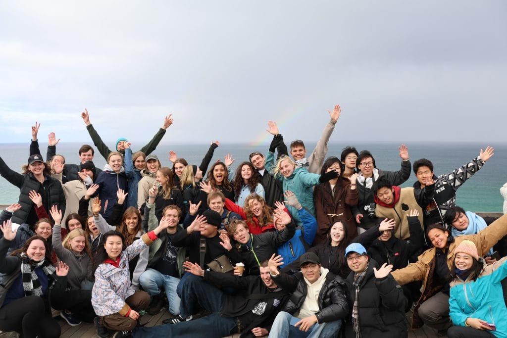 A large group of people pose together outdoors near the ocean, smiling and raising their hands, with cloudy skies and a faint rainbow in the background.