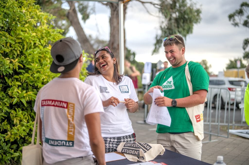 Three people stand outside near a table, talking and laughing. Two wear “Tasmanian University Student” shirts, and one wears a green “The Greens” shirt and holds papers.