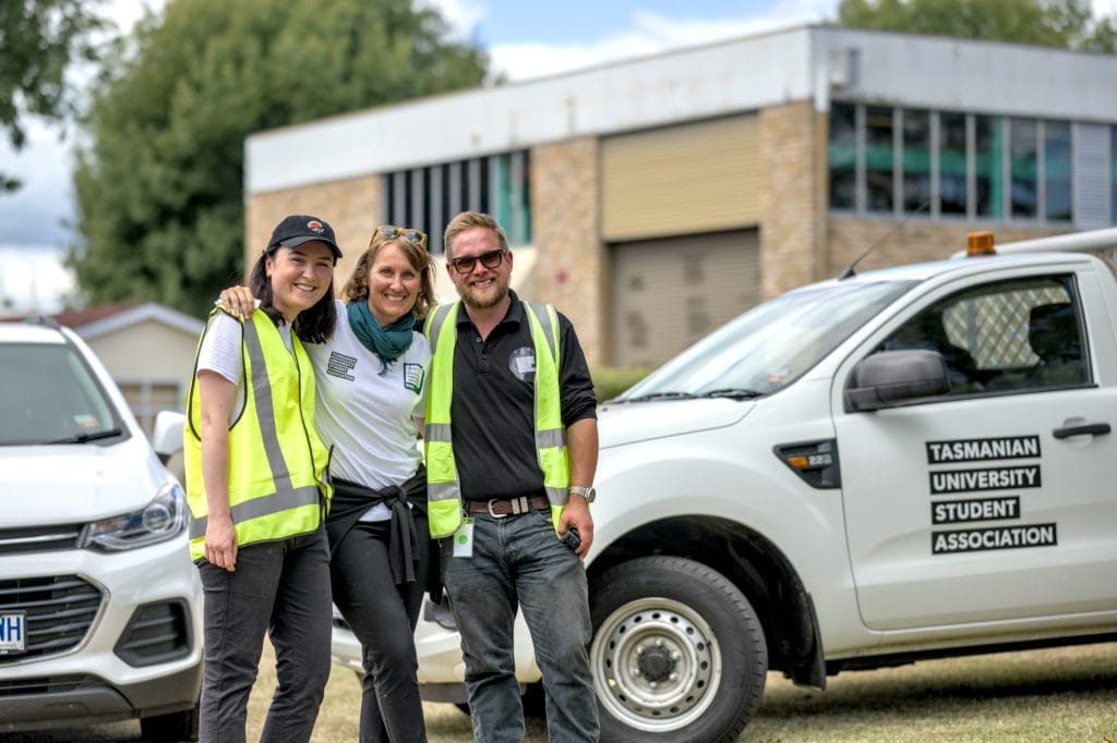 Three people wearing safety vests stand smiling next to a Tasmanian University Student Association vehicle outside a building.