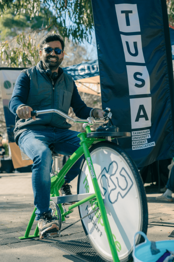 A man wearing sunglasses and a waistcoat sits on a green stationary bike beside a TUSA (Tasmanian University Student Association) banner outdoors.