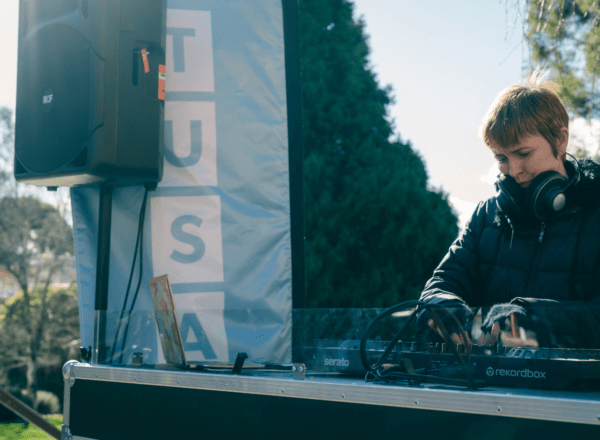 Person wearing headphones operates a DJ controller at an outdoor event, standing beside a speaker and a banner that partially reads "TUSA.