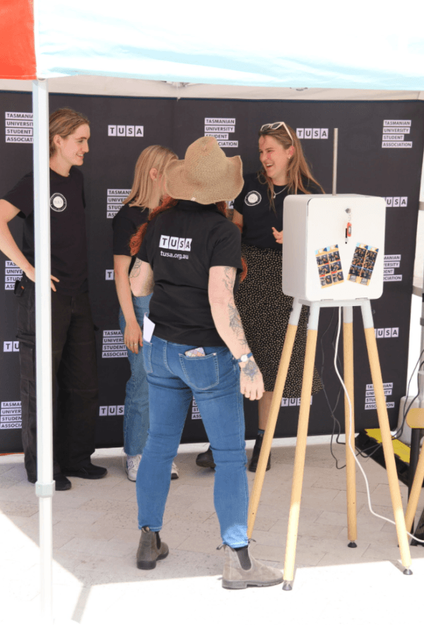 Four people stand and talk in front of a Tasmanian University Student Association backdrop, next to a photo booth under a canopy.