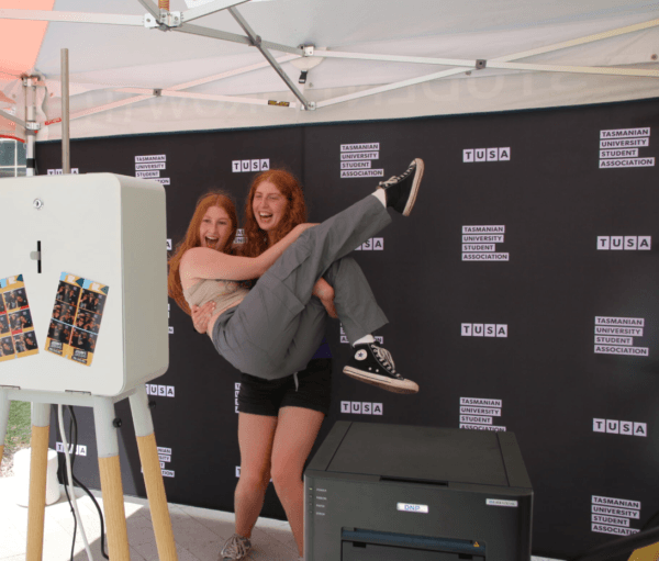 Two people pose for a photo; one lifts the other whilst both smile, standing in front of a Tasmanian University Student Association backdrop at an event stand.