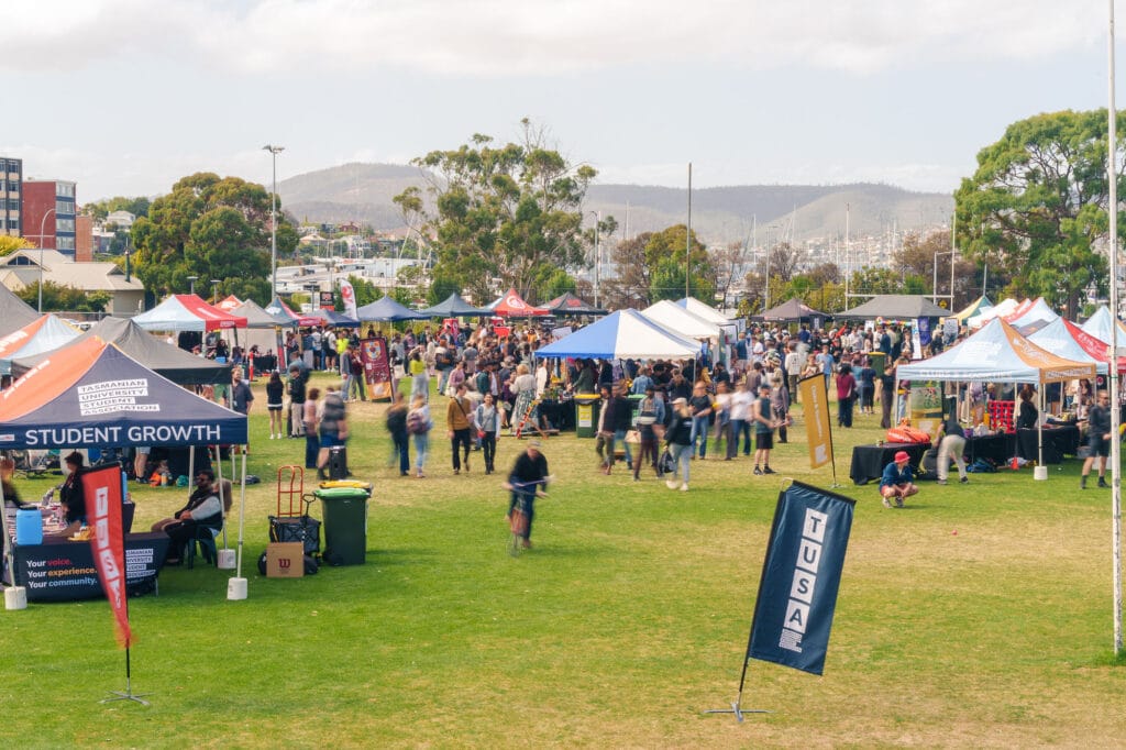 Outdoor event with numerous tents, stalls, and people walking or gathering on a grassy field, with trees and hills visible in the background.