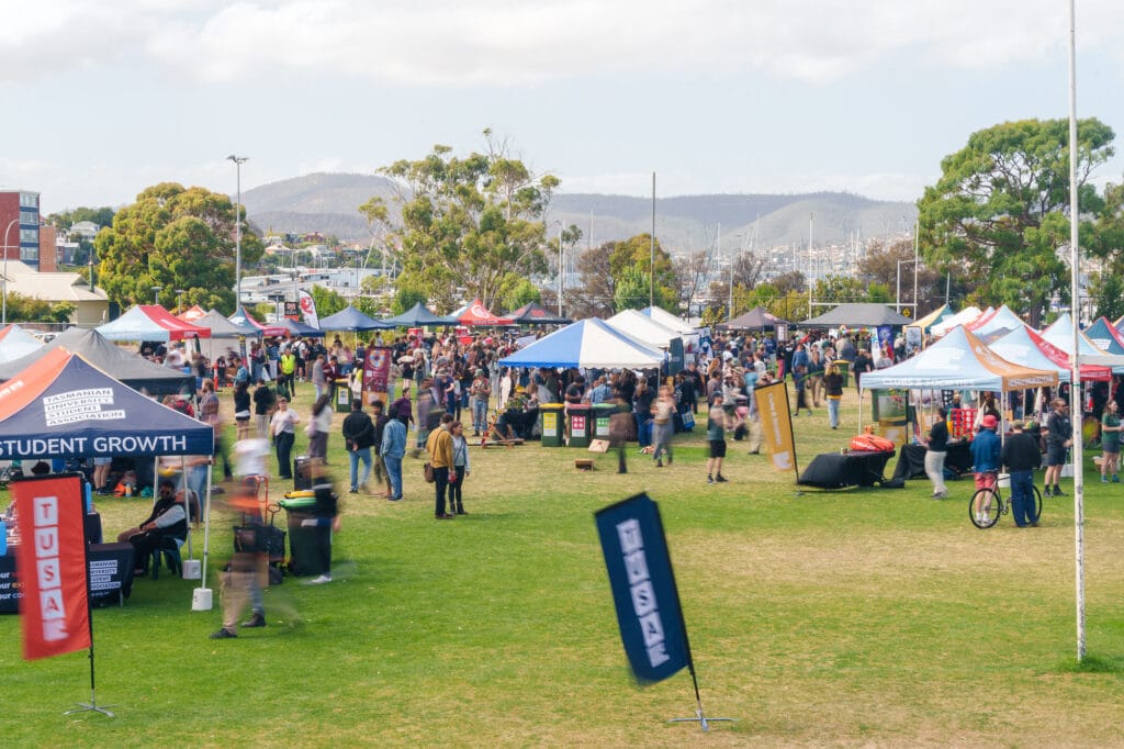 Outdoor event with multiple marquees and stalls, people walking and gathering on a grassy area, trees and hills in the background.