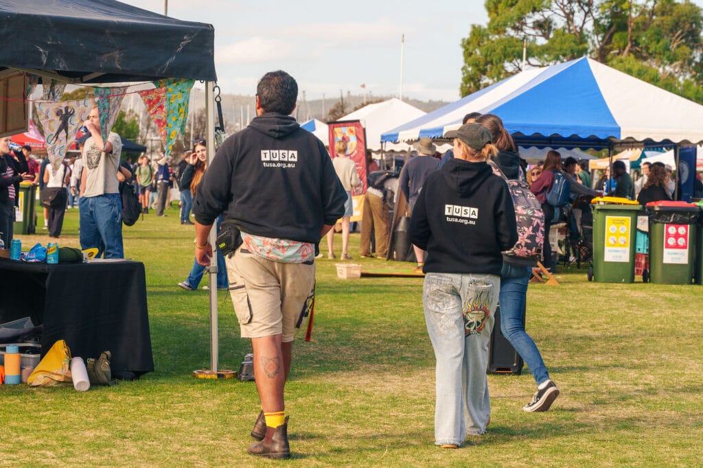 Two people wearing "TUSA" hoodies walk through an outdoor festival with marquees, stalls, and other attendees visible in the background.