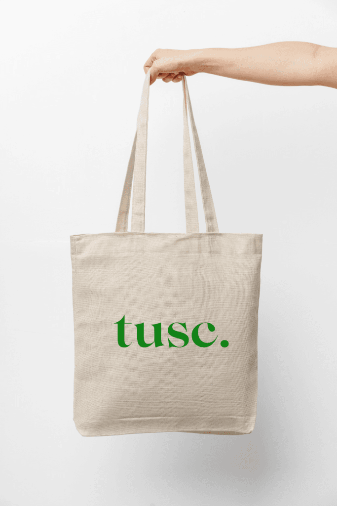 A hand holds a beige canvas tote bag with the word "tusc." printed in green lowercase letters on the front.