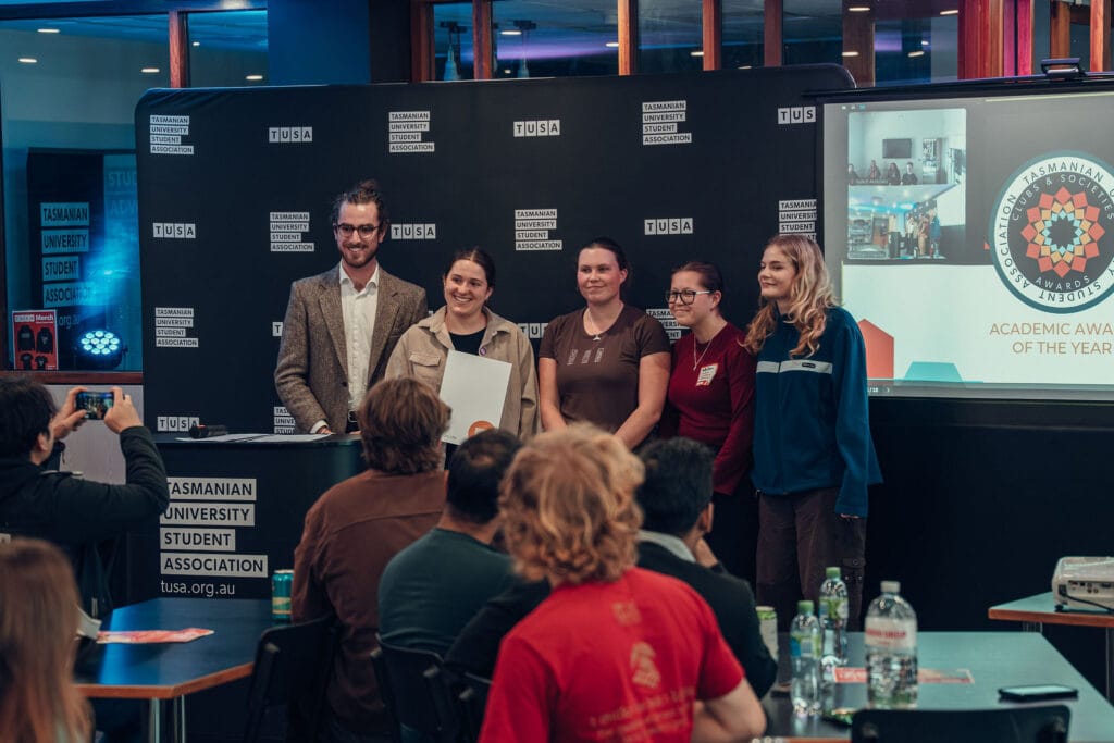 A group of five people stands together on stage at a Tasmanian University Student Association event, posing for a photo whilst others watch and take pictures.